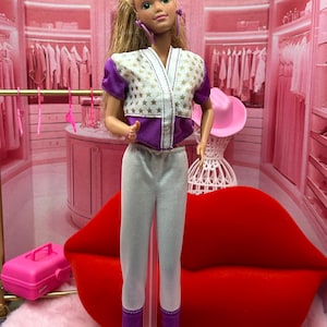 May include: A doll wearing a purple and white star-patterned top, white pants, and white sneakers. The doll has blonde hair and purple earrings. The background is a pink room with a red lip-shaped couch.