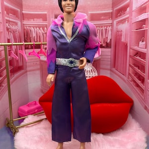 May include: A doll wearing a purple jumpsuit with a silver belt and a star-shaped buckle. The doll has black hair and is standing in a pink room with a red lip-shaped couch and a pink rug.