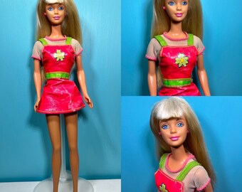 May include: A vintage Barbie doll with blonde hair, blue eyes, and pink lipstick. She is wearing a pink and green dress with a floral design. The doll is standing on a white stand.