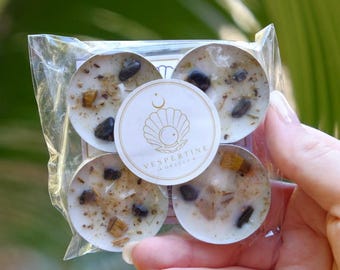 May include: Four white tea light candles packaged in clear plastic, each embedded with small dark stones and dried botanical pieces. A circular white label in the centre features a gold line drawing of a shell and the text VESPERTINE ORACLE.