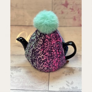 May include: A black teapot with a knitted cosy in shades of purple, pink, and blue. The cosy is topped with a fluffy, light green pom-pom. The teapot is on a light wood-grain surface.