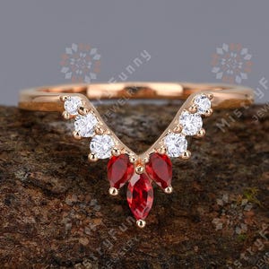 Peut inclure: Une délicate bague en or avec un design en forme de V orné de petites pierres précieuses blanches et de trois pierres précieuses rouges plus grandes.