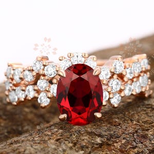 May include: A rose gold ring featuring a large, oval, deep red gemstone. The ring is adorned with smaller, clear gemstones. The band has a floral design with more clear gemstones, creating a sparkling effect.