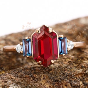 May include: A rose gold ring with a central, rectangular, deep red gemstone, framed by two blue gemstones and smaller white stones. The ring has an intricate design, set against a natural wood background.