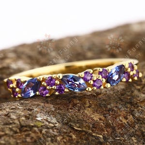 May include: A gold ring with alternating oval blue and round purple gemstones. The ring is displayed on a textured, brown surface, highlighting the jewellery's design and colour contrast.