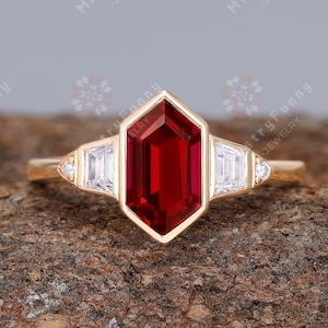 May include: A gold ring featuring a red gemstone and two smaller white gemstones on either side. The ring is displayed on a piece of wood.