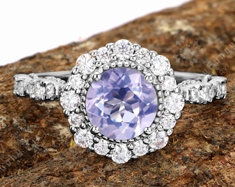 May include: A silver ring featuring a large, round, light purple gemstone surrounded by a halo of smaller white gemstones. The ring's band has a delicate design with additional small white gemstones. The ring is displayed on a piece of brown wood.