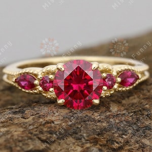 May include: A gold ring featuring a large, round, red gemstone and smaller red gemstones. The ring has an intricate design. The ring is displayed on a textured, brown surface.