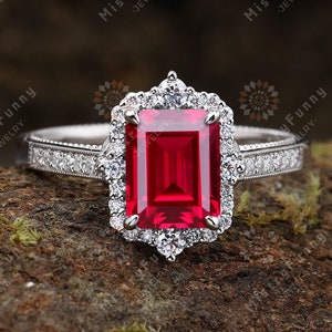 Peut inclure: Une bague en argent avec un gros rubis taille émeraude entouré d'un halo de petits diamants. La bague est posée sur une surface brune et mousseuse.