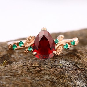 Peut inclure: Une bague en or rose avec une pierre rouge en forme de poire, ornée d'un motif de feuilles délicates et de petites pierres vertes.