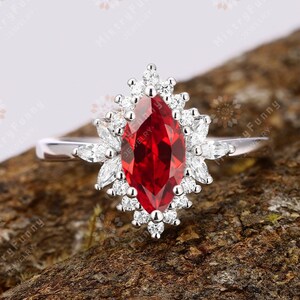 Könnte beinhalten: Ein Ring mit einem roten, marquiseförmigen Edelstein, der von einem Heiligenschein aus kleineren weißen Edelsteinen umgeben ist. Der Ring ist in Silber gefasst und liegt auf einer braunen Holzoberfläche.