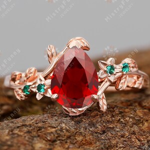 May include: A rose gold ring with a large, pear-cut, deep red gemstone. The ring has floral and leaf detailing, and small green gemstones. The ring is set against a textured, brown background.