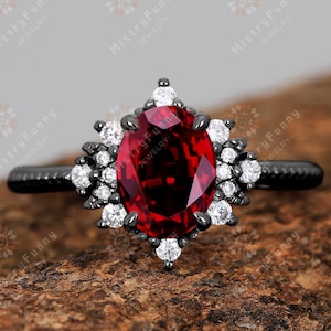 May include: A black ring featuring a large, oval-cut, deep red gemstone. The central stone is encircled by smaller white gems, forming a floral or starburst pattern. The ring's band is black, and the setting is detailed.