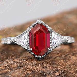 May include: A silver ring with a large, hexagonal, deep red gemstone as the centerpiece. The ring features smaller, triangular white gemstones on either side of the main stone, with intricate detailing along the band.