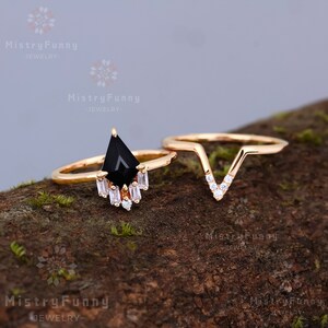 May include: A set of two gold rings with a black gemstone and clear cubic zirconia stones. The larger ring features a black gemstone in a teardrop shape with a row of clear cubic zirconia stones below it. The smaller ring is a V-shape with clear cubic zirconia stones along the edges.