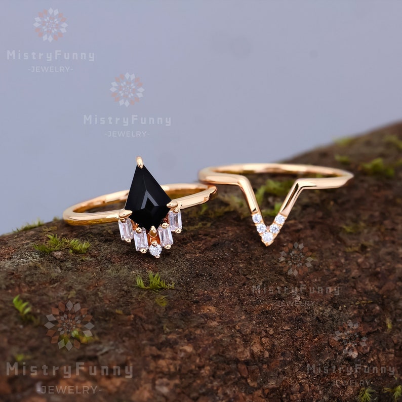 May include: A set of two gold rings with a black gemstone and clear cubic zirconia stones. The larger ring features a black gemstone in a teardrop shape with a row of clear cubic zirconia stones below it. The smaller ring is a V-shape with clear cubic zirconia stones along the edges.