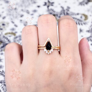 May include: A gold engagement ring set with a black pear-shaped gemstone and a band of smaller white gemstones. The ring is on a finger.