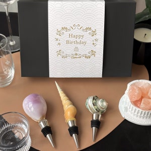 May include: Three decorative bottle stoppers made from seashells. The stoppers are in the shape of a conch shell, a spiral shell, and a clam shell. The stoppers are on a brown surface with a white bowl filled with pink candy.