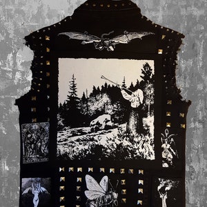 Puede incluir: Chaleco vaquero negro adornado con tachuelas doradas y parches con ilustraciones en blanco y negro. El chaleco tiene cuello y cierre de botón. Los parches incluyen una escena de bosque, una abeja e imágenes góticas.