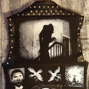 Könnte beinhalten: Schwarze Lederweste mit silbernen Nieten, mit einem weißen und schwarzen Grafikdruck einer Figur auf einer Treppe, zwei schwarzen und weißen Vogelgrafiken, einer schwarzen und weißen Grafik einer Figur in einem Wald und einer weißen Totenkopfgrafik auf einem schwarzen Kreis.