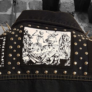 Puede incluir: Chaqueta vaquera negra con tachuelas plateadas y parches. Los parches presentan una imagen en blanco y negro de esqueletos bailando y una imagen en blanco y negro de una mujer con una corona.