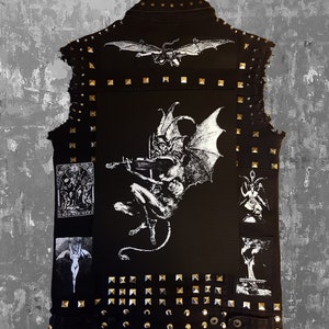 May include: A black denim vest with silver studs and patches. The patches feature a white illustration of a demon playing a violin, a woman with wings, and a scene with multiple figures.