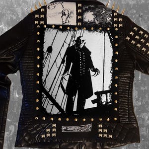 Puede incluir: Chaqueta de cuero negro con tachuelas y púas doradas. La parte trasera presenta un gran gráfico en blanco y negro de una figura en un barco, con parches más pequeños arriba y abajo. La chaqueta tiene un diseño acolchado en los hombros.