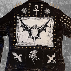 Op de afbeelding: Zwart leren jack met gouden studs en een Ankh-symbool op de rug. Het jack heeft een zwart-witte grafische print van een vleermuis, een skelet en twee vogels.