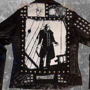 Könnte beinhalten: Schwarze Lederjacke mit silbernen Nieten und einem grafischen Druck eines Mannes in einem schwarzen Anzug, der auf einem Schiff steht. Die Grafik ist in Schwarzweiß.