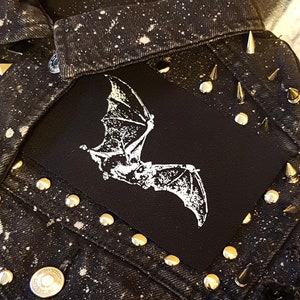 May include: A black denim jacket with white splatter paint and silver studs. A black patch with a white bat design is sewn onto the jacket.