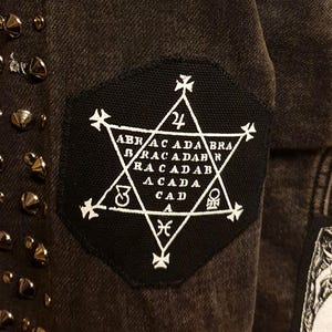 Könnte beinhalten: Eine dunkelgraue Jeansjacke mit silberfarbenen, pyramidenförmigen Metallnieten in vertikalen Reihen. Ein schwarzer, sechseckiger Aufnäher ist auf den Stoff genäht und zeigt einen weißen sechszackigen Stern mit dem Text ABRACADABRA und Symbolen.