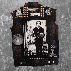 Op de afbeelding: Een zwart denim vest met een skelet en een man die een schedel vasthoudt, bedrukt op de voorkant. Het vest heeft zilveren studs en patches.