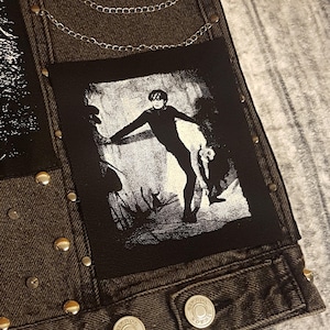 Könnte beinhalten: Dunkelgraue Jeansjacke mit silbernen Nieten und einer silbernen Kette. Die Jacke hat zwei schwarz-weiße Aufnäher mit grafischen Illustrationen. Die Jacke hat zwei silberne Knöpfe mit dem Text "BIKBOY".