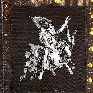 Peut inclure: Écusson carré noir avec une illustration blanche d'une figure chevauchant une créature à cornes. La figure a de longs cheveux et lève un bras. L'écusson est sur une veste en jean noire avec des clous dorés.