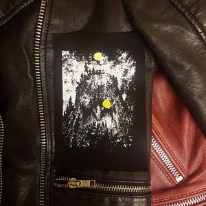 Pode incluir: Patch preto e branco com uma cena de floresta e dois círculos amarelos. O patch é costurado em uma jaqueta de couro preta.