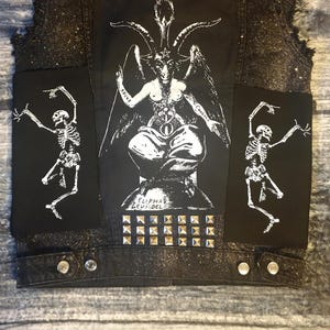 May include: A black denim vest with a white design of a goat-headed figure with wings and the text "Solve" and "Coagula" on the back. The vest has two black patches with white skeleton figures on the front and a row of silver studs along the bottom.