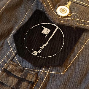 Op de afbeelding: Een donkergrijs spijkerjack met een zwarte stoffen patch op de borstzak. De patch toont een witte cirkelvormige afbeelding met een verticale lijn en een klein vlagpictogram. Het jack heeft zichtbare lichtgekleurde stiksels en metalen knopen.