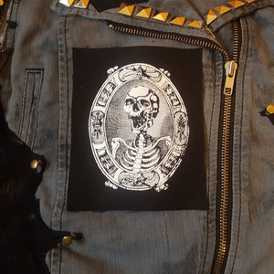 May include: A black and white embroidered patch featuring a skeleton in a circle with ornate details. The patch is sewn onto a gray denim jacket.