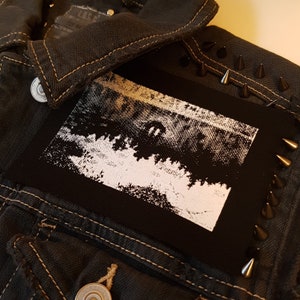 May include: Black denim jacket with a white patch sewn on the front. The patch features a black and white image of a forest scene. The jacket has silver studs along the seams.