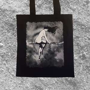 May include: Black tote bag with a white graphic print of a woman riding a broomstick in the clouds.