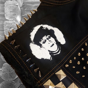 May include: A black denim jacket with silver studs and spikes. A white patch with a black and white image of a person wearing a fur hat is sewn onto the jacket.