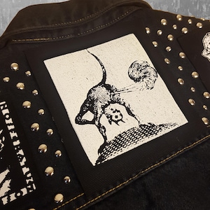 May include: Black and white patch featuring a graphic of a creature with a tail, standing on its hind legs, with its head tilted back and its mouth open. The creature is emitting a cloud of smoke from its rear end.