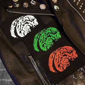 Op de afbeelding: Drie zwarte stoffen patches met hersenontwerpen in wit, groen en oranje worden getoond op een zwarte leren jas. De jas heeft een rits en metalen studs. Bovenop zijn verschillende speldjes zichtbaar.