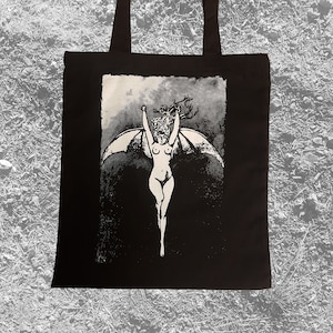 May include: A black tote bag with a white graphic print of a woman with wings spread wide. The woman is nude and has long hair. The background is black with a white cloud.