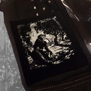 Può includere: Tessuto nero con una toppa quadrata con un'illustrazione in bianco e nero di una figura in un ambiente forestale. Borchie argentate sono visibili vicino al bordo superiore. L'articolo sembra far parte di una giacca o di un gilet.