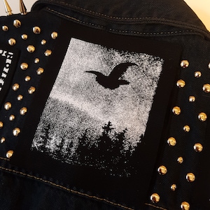 Op de afbeelding: Een zwart denim jack met zilveren studs en een witte patch met een silhouet van een vogel die over een bos vliegt.