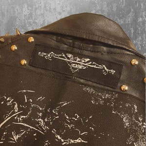 May include: Close-up of a black leather jacket with gold studs and a patch featuring a sword and emblem design. The jacket has a canvas-like material on the lower back with a white graphic print.