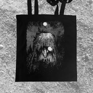 May include: Black tote bag featuring a detailed, grayscale illustration of a creature with a shaggy, textured appearance, set against a dark forest backdrop. A bright, circular moon and a glowing eye-like detail add contrast. The bag has black handles.