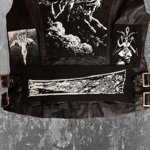 Könnte beinhalten: Schwarze Lederweste mit mehreren weißen Patches mit grafischen Designs. Die Weste hat einen schwarzen Stoffgürtel mit einem weißen grafischen Design.