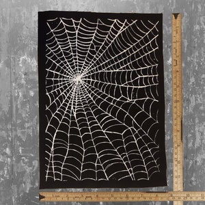 K&ouml;nnte beinhalten: Schwarzer Stoff-Patch mit einem wei&szlig;en Spinnennetz-Design. Das Netz ist detailliert und filigran und strahlt von der Mitte aus.